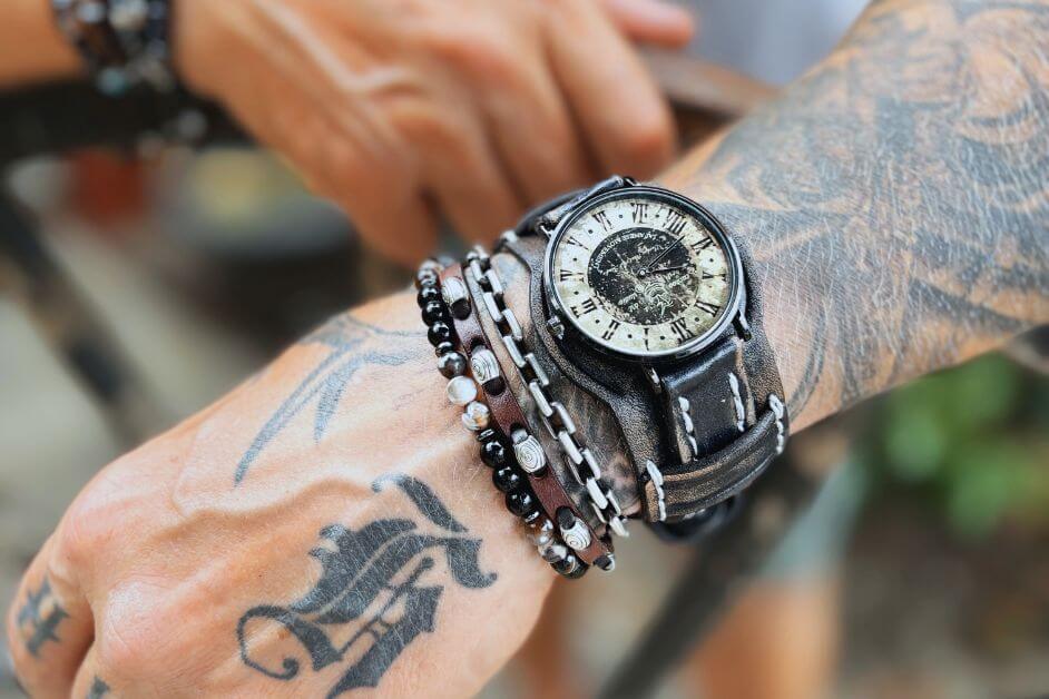 Men's watch with leather band styled with stacked bracelets on tattooed wrist

